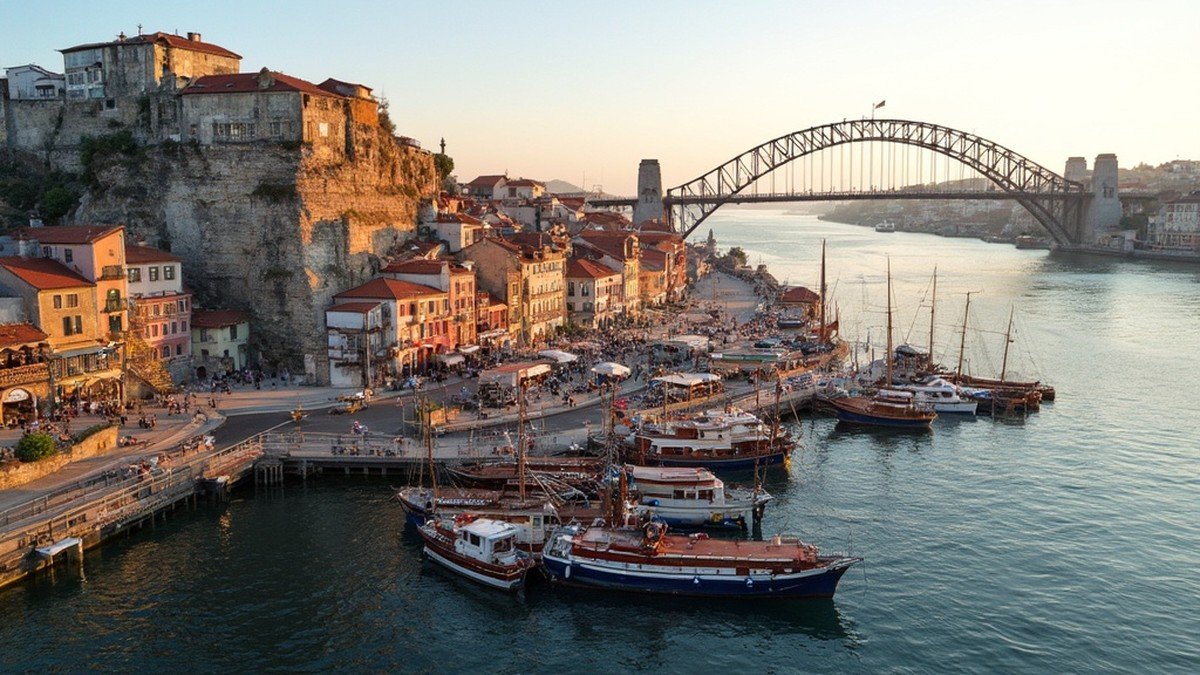 Ribeira riverfront of stacked colourful houses on the Douro with traditional rabelo port boats moored below the iron Ponte Luis I bridge at golden hour, Porto