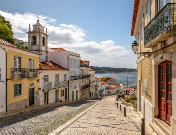 A scenic view of Setúbal, Portugal, showcasing the Sado River, Arrábida Natural Park mountains, and traditional boats docked along the waterfront under a sunny sky.